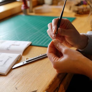 Peut inclure: Un stylet fin en métal noir tenu à la main, positionné au-dessus d'un petit objet. À proximité, sur la surface en bois, se trouvent un livret d'instructions ouvert avec des schémas et du texte, un cutter en métal argenté et un tapis de découpe vert.