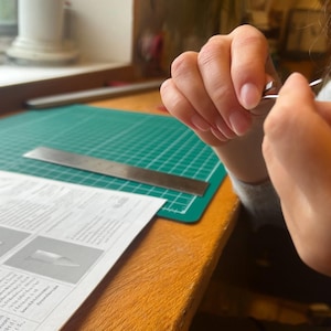 May include: A silver-colored metal component held between fingers, positioned near a white instructional paper with diagrams, a metal ruler, and a green cutting mat on a wooden surface, showing a small, pointed, and curved metallic object.