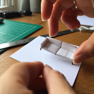 May include: A miniature paper sofa model featuring a white surface with thin black outlines, constructed from folded cardstock. The small-scale furniture piece displays a rectangular seat and backrest design, showcasing precise papercraft assembly techniques.