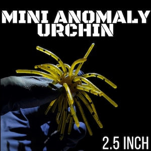 May include: A yellow, multi-legged fishing lure with black speckles, labeled as a 2.5 inch Mini Anomaly Urchin. The lure features a central body with numerous thin, radiating appendages extending outward against a solid black background.