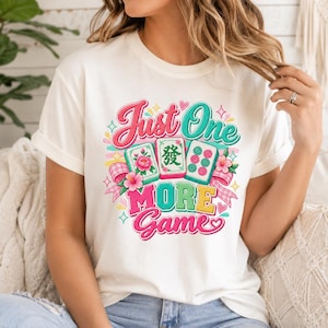 May include: A cream-coloured t-shirt featuring a colourful graphic print with three mahjong tiles, pink hibiscus flowers, and a gingham ribbon. The text reads Just One MORE Game in a mix of pink, teal, and yellow script and block lettering.
