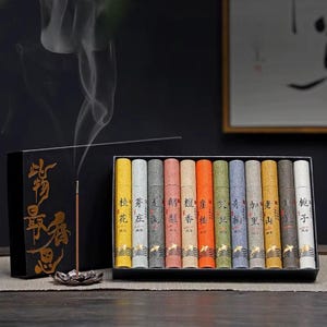 May include: A set of twelve cylindrical incense tubes in a black box, each featuring distinct colors and Chinese characters. A single incense stick burns in a metallic lotus-shaped holder, emitting a thin trail of smoke beside the box.