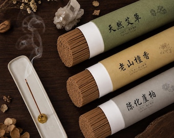 May include: Three cylindrical cardboard tubes containing brown incense sticks, each with distinct Chinese text and botanical illustrations. A single incense stick burns in a white ceramic holder, emitting a thin trail of smoke.