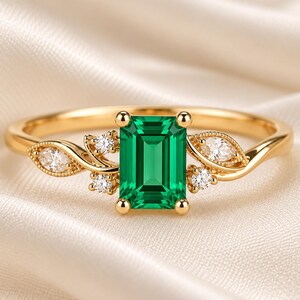 Puede incluir: Un anillo de oro con una piedra preciosa verde de corte esmeralda sujeta por cuatro garras. La banda está adornada a ambos lados con una piedra clara de corte marquesa y dos pequeñas piedras claras redondas, creando un diseño simétrico.