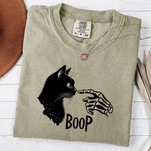 May include: A light olive green t-shirt featuring a black graphic of a cat profile and a skeleton hand touching its nose. The text BOOP is printed below the graphic. The inner neck label displays the Comfort Colors brand name and size S P CH.