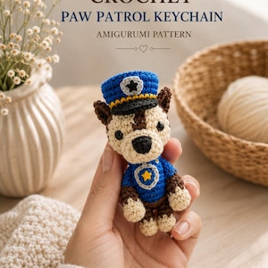 Può includere: Una piccola figura amigurumi all'uncinetto che rappresenta un cane con un'uniforme da polizia blu e un berretto abbinato con visiera nera ed emblema a stella. La figura presenta dettagli in filato marrone e crema, occhi neri e gilet blu con stella.