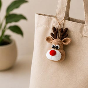 Può includere: Una borsa tote in tela beige con un ornamento a forma di renna lavorato all'uncinetto attaccato al manico. La renna presenta corna marrone scuro, orecchie marrone chiaro, muso bianco, grandi occhi bianchi e neri e un naso rosso.