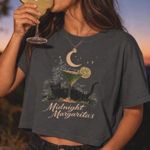 May include: A charcoal gray short-sleeve t-shirt featuring a graphic print of a black cat lying near a green margarita glass with a lime slice, a crescent moon, stars, and the text Midnight Margaritas in a light-colored script font.