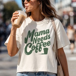 Könnte beinhalten: Ein cremefarbenes Kurzarm-T-Shirt mit dem dunkelgrünen Schriftzug Mama Needs Coffee in einer fetten, wellenförmigen Schriftart. Das Design enthält eine kleine Grafik einer Kaffeetasse neben dem Text. Das T-Shirt hat einen Rundhalsausschnitt.