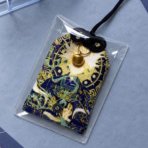 May include: A rectangular fabric omamori charm featuring intricate embroidery in shades of deep blue, gold, and white. The charm is encased in a clear plastic sleeve, topped with a black cord and a small gold-toned metal bell attached to the top.