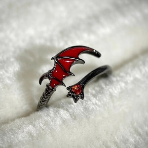 May include: A black metal ring featuring a stylized bat wing design filled with vibrant red enamel. The band terminates in a pointed claw-like detail that holds a small, round, faceted red gemstone, creating a cohesive dark aesthetic.