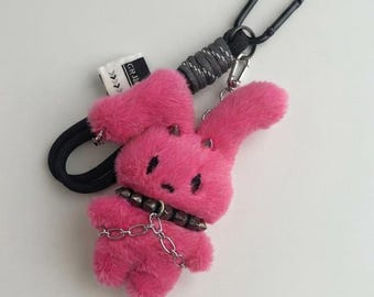 May include: A bright pink plush rabbit keychain featuring a black studded collar and a silver-toned metal chain draped across its chest. The rabbit has two small horns on its head and is attached to a black carabiner clip with a black cord.
