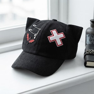 May include: A black baseball cap featuring two pointed cat-like ears on the crown. The front panel displays a red and black bat patch attached to silver safety pins with a small chain, and a white cross patch with a red border on the side.