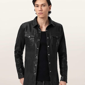 Puede incluir: Una camisa negra de manga larga con botones, hecha de un material liso, con cuello en punta, dos bolsillos en el pecho con solapas abotonadas y botones de presión metálicos de color plateado en la parte frontal y en los puños.