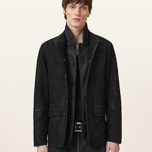 May include: A black suede blazer featuring a notched lapel, a button-front closure, and two flap pockets at the waist. The jacket is worn over a dark gray shirt and is paired with dark gray denim jeans featuring a black leather belt with a silver buckle.