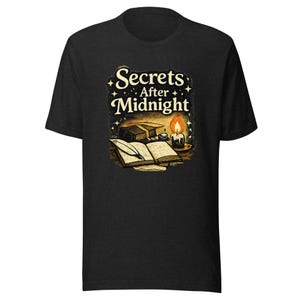 Può includere: Una t-shirt nera a maniche corte con una stampa grafica raffigurante un libro aperto, una penna d'oca e una candela accesa. Il testo "Secrets After Midnight" è stampato in color crema sopra l'illustrazione, circondato da piccole icone a stella.
