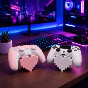 Puede incluir: Dos soportes para mandos de juego con forma de corazones pixelados, uno en rosa claro y otro en blanco. El soporte rosa sostiene un mando a juego en rosa claro y blanco, mientras que el blanco sostiene un mando blanco y negro con botones.