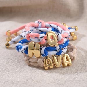 May include: A collection of three braided cord bracelets featuring gold-toned metal letter charms. One bracelet is pink, one is blue, and one is beige. The charms spell out the letters Q, N, A, V, and A, with gold-toned metal beads at the ends.