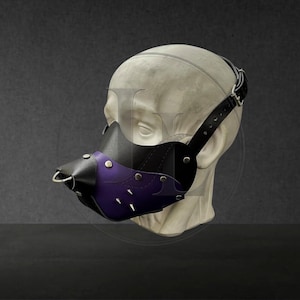 May include: A black and purple face mask featuring a pointed snout design, silver-toned metal studs, and a small metal ring at the tip. The mask is secured with adjustable black straps and metal buckles, fitted onto a neutral-toned mannequin head.