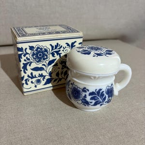 Op de afbeelding: Een kleine scheerkom van wit melkglas met bijpassend deksel, voorzien van een blauw bloemmotief. De kom heeft één handvat en staat naast de originele vierkante kartonnen doos, die is versierd met een bijpassend blauw bloemmotief.