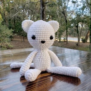 May include: A white crocheted stuffed animal featuring a round head with two small ears, a small torso, and long limbs. The creature has two large, round, black plastic eyes and a small, triangular black plastic nose centered on its face.