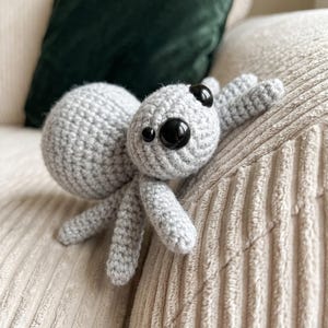 May include: A light grey crocheted spider toy featuring a round body and head with two large, shiny black plastic eyes and two smaller black eyes positioned above them. The spider has multiple segmented legs extending from its central body.