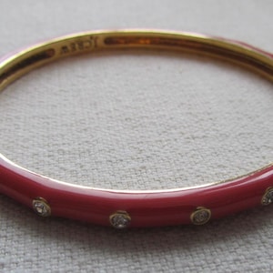 Op de afbeelding: Een goudkleurige armband met een rode emaille afwerking. De armband heeft kleine, heldere, ronde edelstenen die in het rode emaille zijn gezet. De binnenkant van de armband is goudkleurig en de merknaam "J.Crew" is zichtbaar.