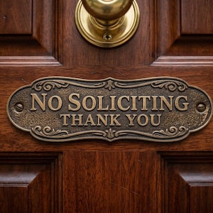 Könnte beinhalten: Ein ovales, bronzefarbenes Metallschild mit dekorativen Schnörkeln an den Rändern, das an einer dunklen Holztür befestigt ist. Das Schild trägt die erhabene Aufschrift NO SOLICITING THANK YOU und ist mit zwei sichtbaren Schrauben befestigt.