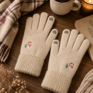 Könnte beinhalten: Ein Paar beigefarbene Strickhandschuhe mit einer kleinen Stickerei eines Schneemanns und eines grünen Baumes auf dem Handrücken. Die Handschuhe haben gerippte Bündchen und graue, für Touchscreens geeignete Fingerspitzen an Daumen und Zeigefinger.