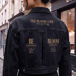 May include: A black denim jacket featuring gold text on the back. The text reads THE BLOOM CODE COCOA & BLOOM EST. 2026, BE YOURSELF intelligent stylish cultured, and BLOOM YOUR WAY expressive confident unapologetic. The jacket has a collar and buttons.