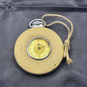 May include: A round, tan-colored pocket watch with a yellow face featuring black numerals and hands. The watch has a silver-toned metal crown at the top and a short, beige-colored rope cord attached to the crown for hanging or carrying purposes.