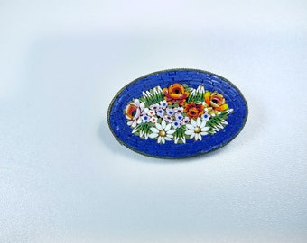 Peut inclure: Broche ovale en mosaïque avec un motif floral. La broche présente un fond bleu profond avec un arrangement détaillé de fleurs colorées, dont des roses et des marguerites. La mosaïque est encadrée d'une fine bordure métallique.
