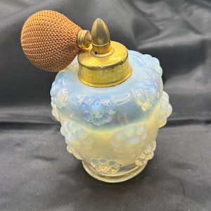 May include: An opalescent glass perfume bottle with a raised floral pattern, featuring a gold-toned metal atomizer cap and a textured, round, tan-colored fabric bulb. The glass body displays a milky, iridescent finish with translucent blue and white tones.