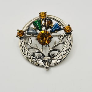 May include: A silver-toned brooch with a deer head design. The brooch features a central amber-colored gemstone surrounded by smaller gemstones in amber, green, brown, gray, and blue. The brooch has an intricate Celtic knot design.