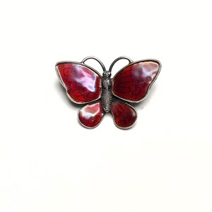 May include: A silver-toned butterfly brooch with red enamel wings. The brooch has a detailed design, with the red enamel wings outlined in silver. The butterfly's body is also silver, and the antennae are curved.