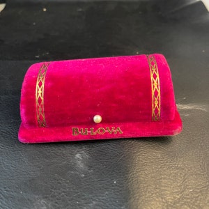 Puede incluir: Una caja de joyería de terciopelo rojo con ribetes dorados y la palabra "BULOVA" impresa en la parte delantera. La caja tiene un botón blanco perlado en la parte superior.