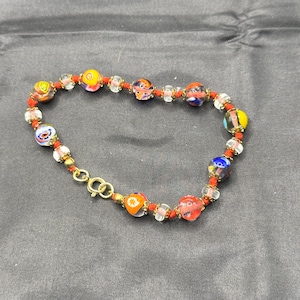 May include: A bracelet featuring multicolored glass beads with intricate patterns, interspersed with smaller clear glass beads and gold-toned metal spacers. The beads are strung on a thin red cord and secured with a gold-toned metal spring ring clasp.