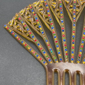 May include: A brown plastic hair comb with a fan-shaped design. The comb has a row of colorful rhinestones along each tooth. The rhinestones are red, blue, green, and yellow.