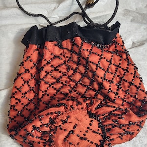 Puede incluir: Un bolso con cordón naranja con cuentas, con la parte superior y el cordón negros. La bolsa está cubierta con un patrón de rejilla de cuentas negras. El cordón es un cordón marrón oscuro con una cuenta dorada decorativa.