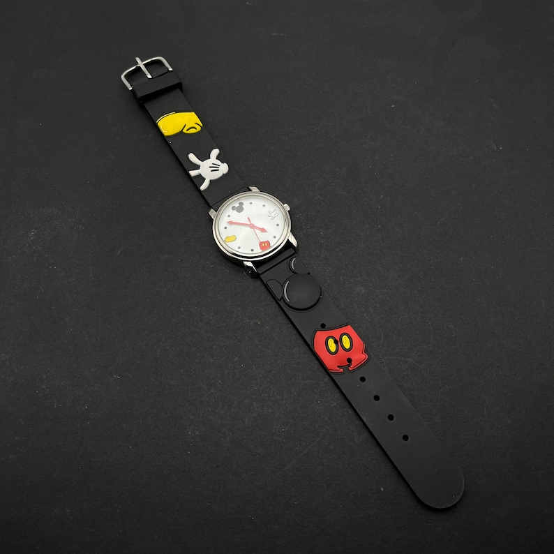 May include: Black wristwatch with a white face and Mickey Mouse characters on the band. The watch has a silver buckle.