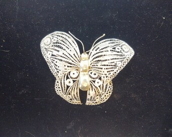 May include: A silver butterfly brooch with intricate filigree detailing. The wings feature delicate openwork patterns, and the body has a smooth, polished finish. The brooch is set against a dark background.