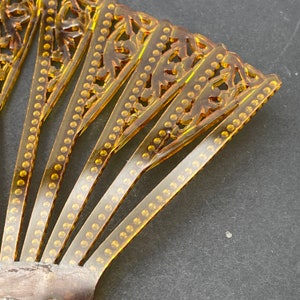 May include: A gold-toned plastic hair comb with a decorative, fan-shaped design. The comb has a series of teeth and a row of small, round embellishments along each tooth.