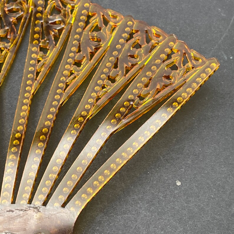 May include: A gold-toned plastic hair comb with a decorative, fan-shaped design. The comb has a series of teeth and a row of small, round embellishments along each tooth.