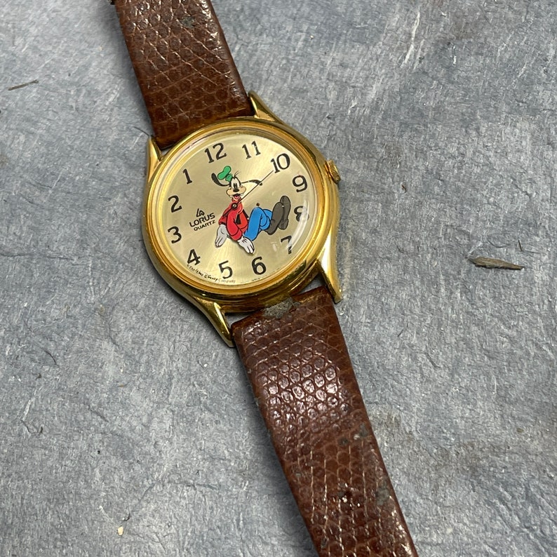 Pu&ograve; includere: Un orologio da polso color oro con un cinturino in pelle marrone. Il quadrante dell'orologio presenta un'immagine di cartone animato di Paperino, il testo "Lorus Quartz" e i numeri da 1 a 12.