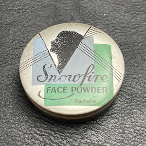 May include: A vintage round metal compact with a silver lid and a black and white silhouette of a woman's face. The lid has a green and blue geometric design and the text "Snowfire Face Powder Rachelle".