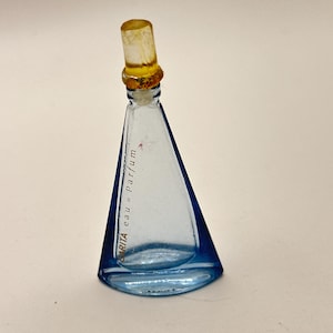 Puede incluir: Un pequeño frasco de perfume triangular con un cuerpo de vidrio azul y un tapón de color amarillo. El frasco tiene una etiqueta con el texto "Rita eau de Parfum" en una fuente cursiva. El frasco está vacío y parece vintage.
