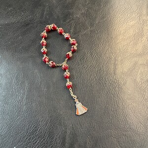 May include: A single-decade rosary featuring faceted deep red glass beads, each encased in ornate silver-toned metal caps. The beads are connected by a silver-toned metal chain, terminating in a small, triangular silver-toned metal pendant charm.