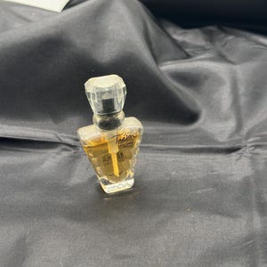 Puede incluir: Un pequeño frasco de perfume transparente con un tapón facetado. El frasco contiene un líquido dorado y está etiquetado como "Trésor" de Lancôme. El frasco se encuentra sobre un fondo oscuro y texturizado.