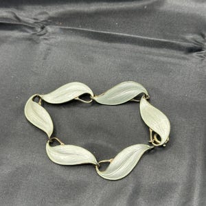 Puede incluir: Una pulsera plateada con diseño de hojas. Cada eslabón en forma de hoja tiene una superficie texturizada y está conectado por pequeños bucles dorados. La pulsera se muestra sobre un fondo oscuro.