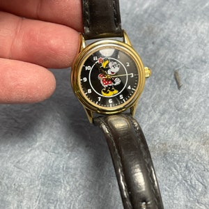 May include: A gold-coloured wristwatch with a black face featuring a Minnie Mouse graphic. The watch has a black leather strap and gold-coloured hands and hour markers. The watch face is approximately 2.5 cm in diameter.